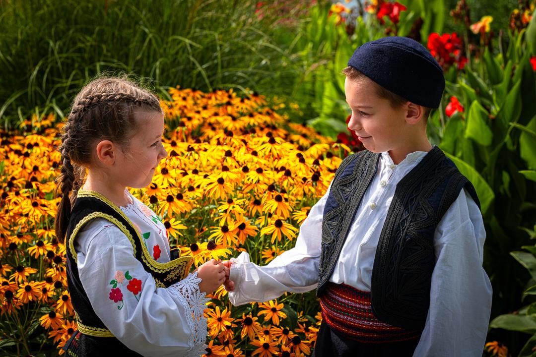 Ein Mädchen und ein Junge der Teslići in Tracht reichen sich zwischen Sonnenhut-Blumen die Hand